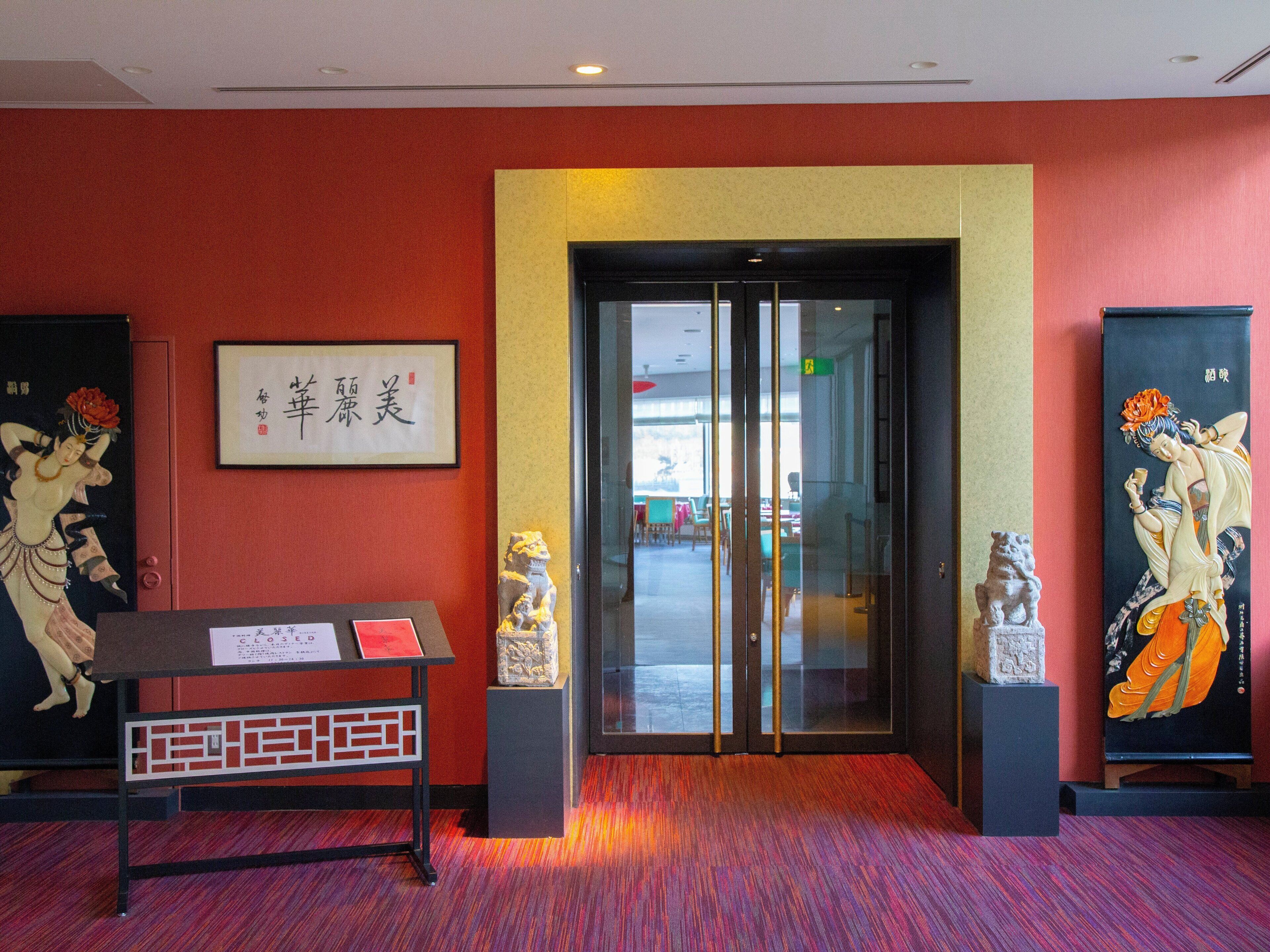 A hotel lobby with a red carpet, a reception desk, and two statues on either side of a glass door.