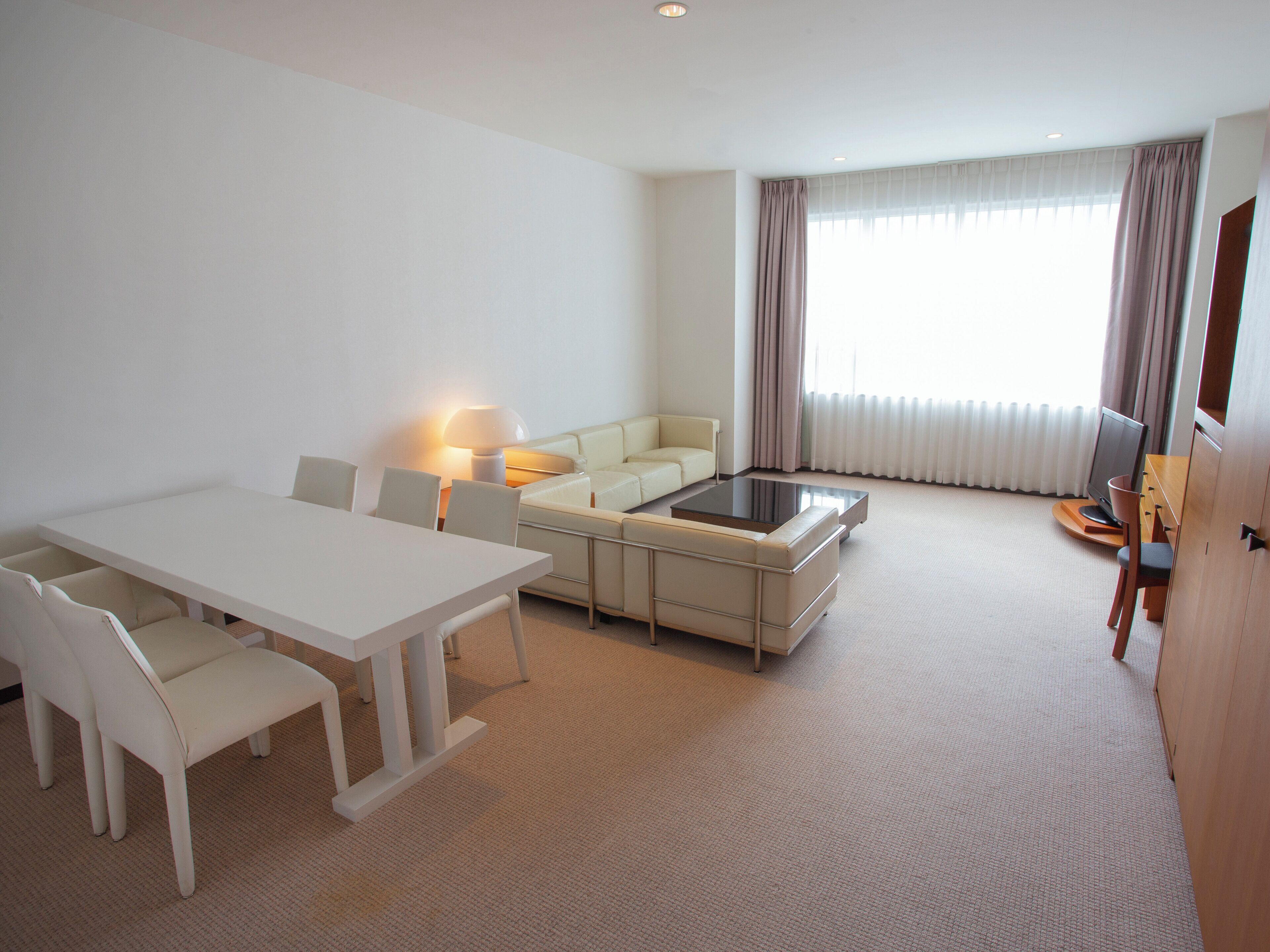A modern hotel room with a dining area, a sofa, a TV, and a window with curtains.