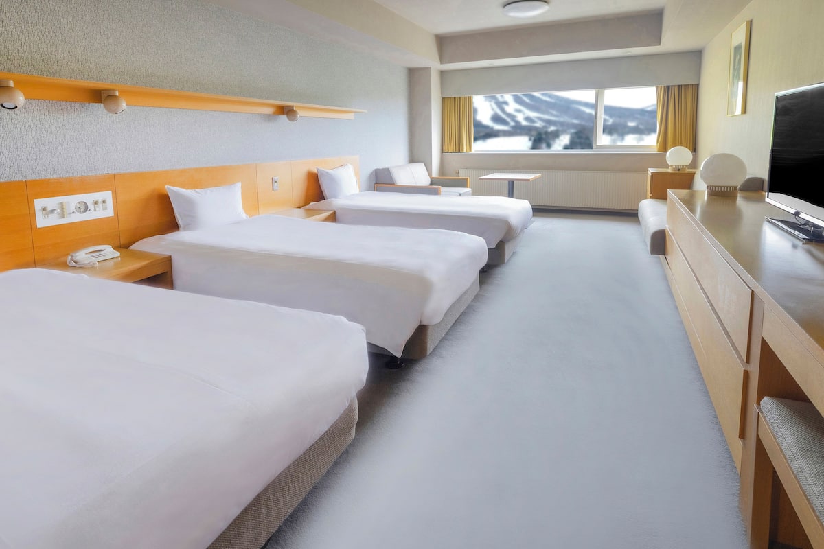 A hotel room with three beds, a desk, a TV, and a window with a view of mountains.