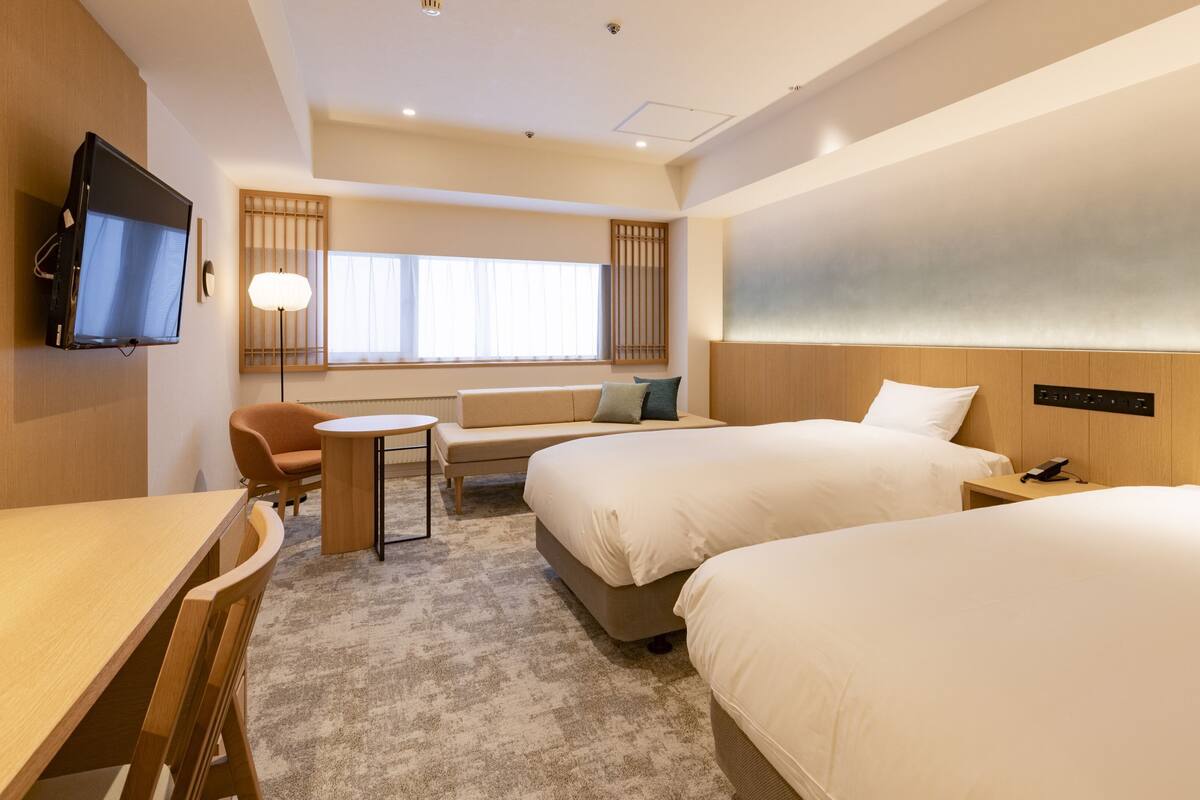 A hotel room with two beds, a TV, a desk, a chair, a sofa, and a window with curtains.