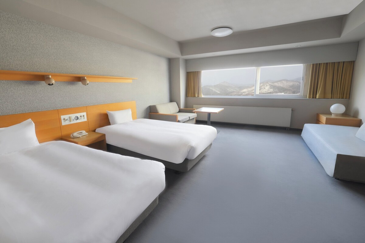 A hotel room with two beds, a small table, a chair, and a window with a mountain view.