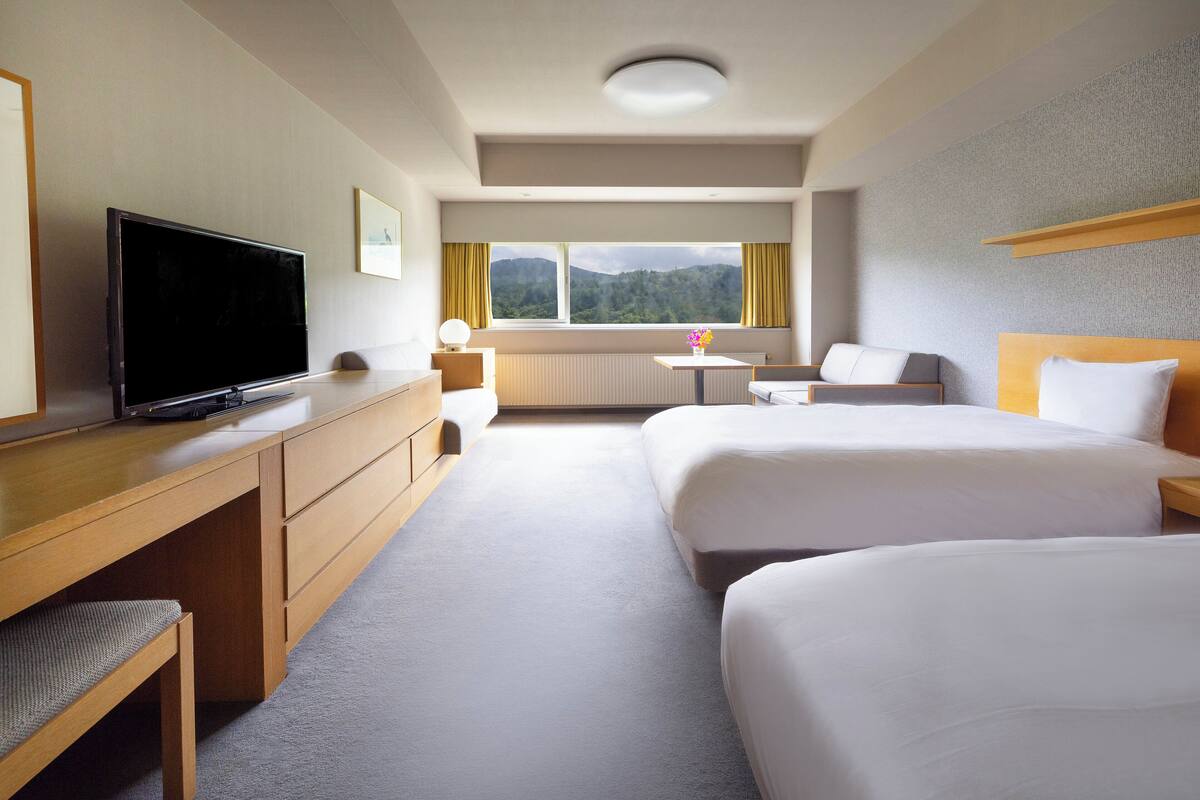A hotel room with two beds, a desk with a TV, a small table, and a view of mountains through the window.