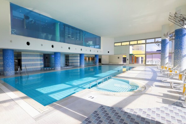 Una amplia piscina cubierta con techo de cristal, suelo de azulejos azules y sillones de descanso blancos.