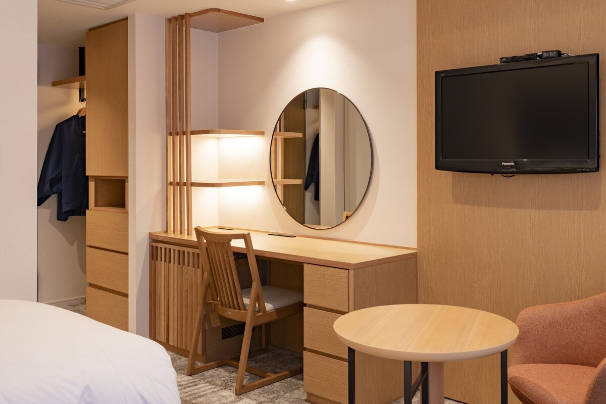 Habitación de hotel con escritorio de madera, espejo redondo, televisor, cama y una silla.