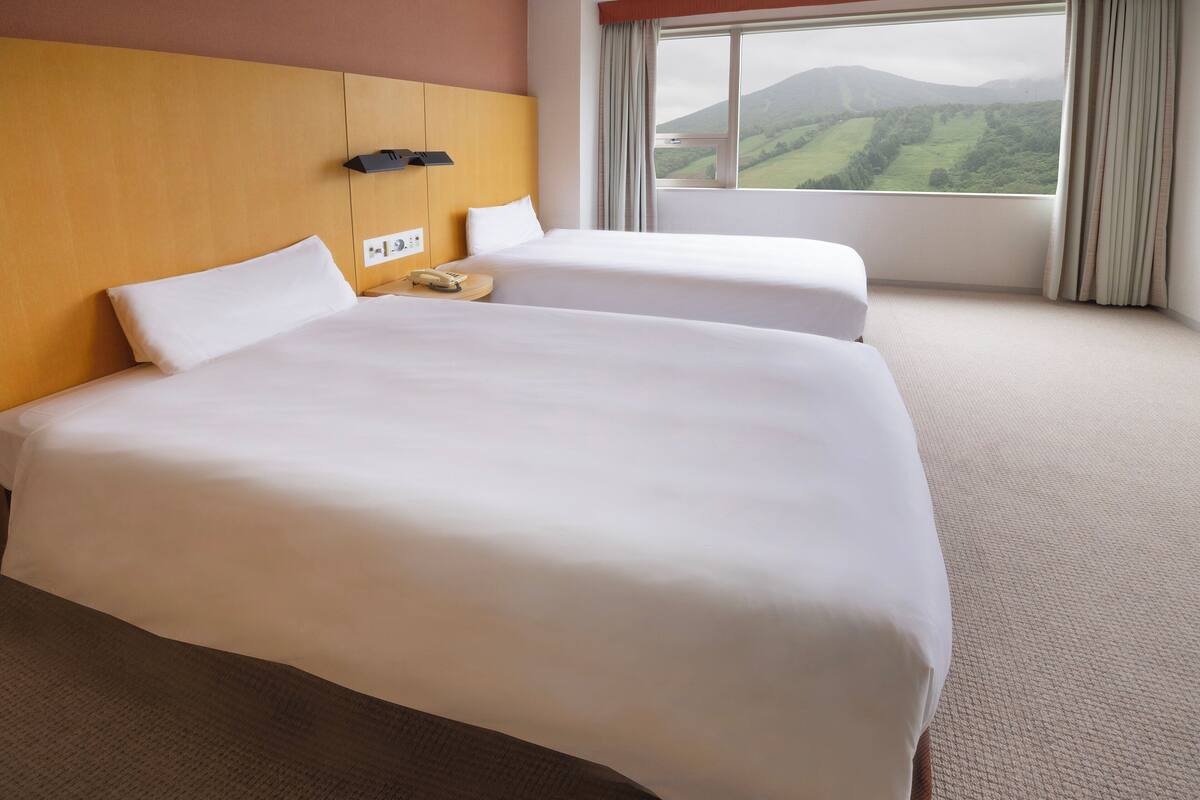 A hotel room with two beds, a wooden headboard, a window with a view of green hills, and a small table with a telephone.