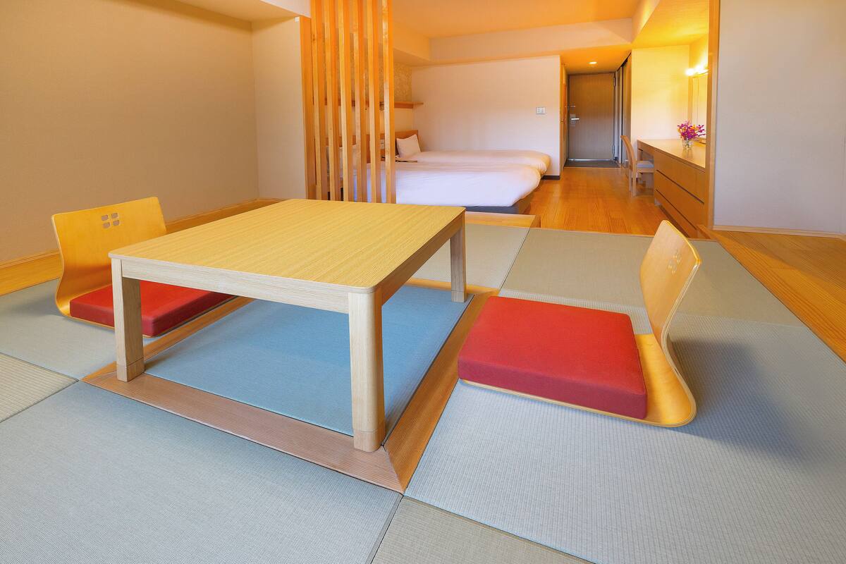 Una habitación con una mesa y sillas de madera, una cama y un espejo con un jarrón de flores.