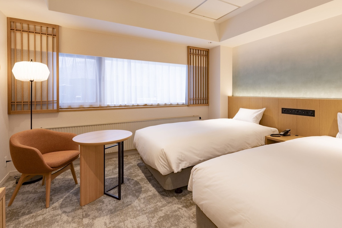 A hotel room with two beds, a small table, a chair, a lamp, and a large window with blinds.