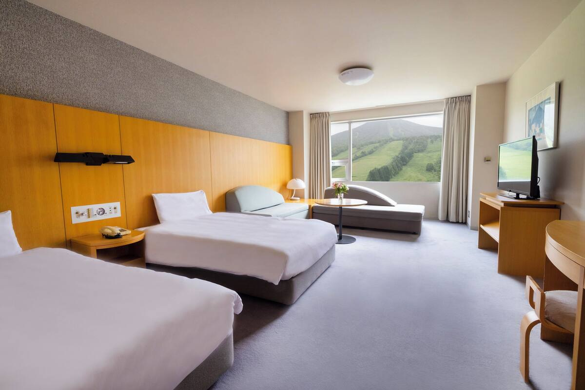 Habitación de hotel con dos camas, un sofá, un escritorio, un televisor y un amplio ventanal con vistas al campo.