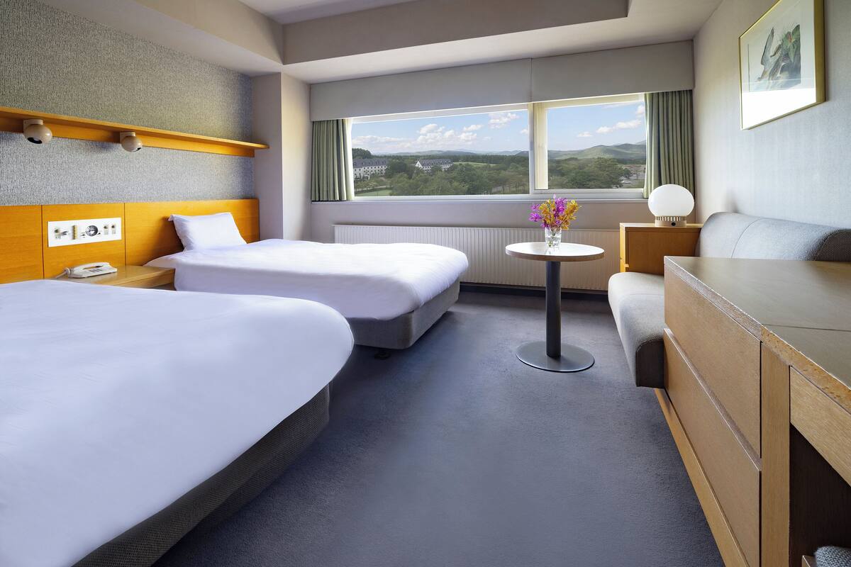A hotel room with two beds, a desk, a chair, a small table with flowers, and a window with a view of greenery and hills.