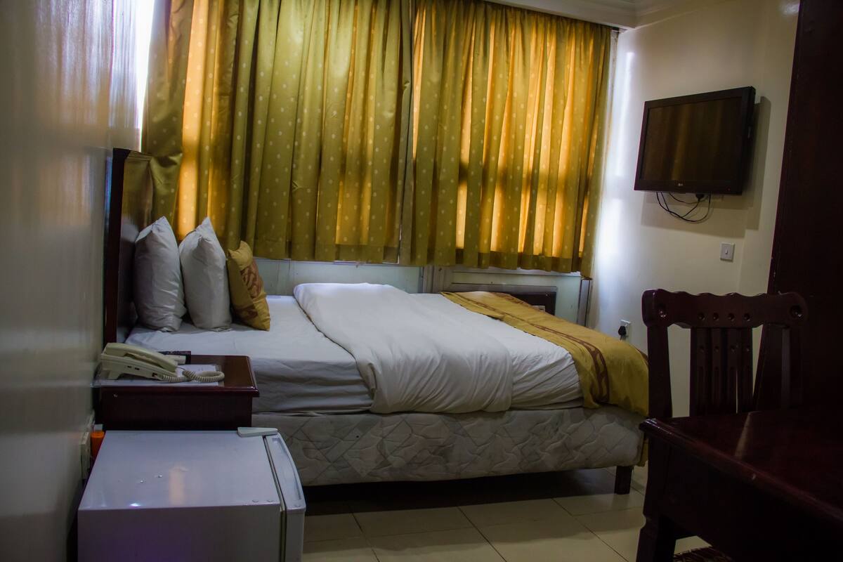 A hotel room with a bed, a TV mounted on the wall, a wooden chair, and a desk with a telephone.