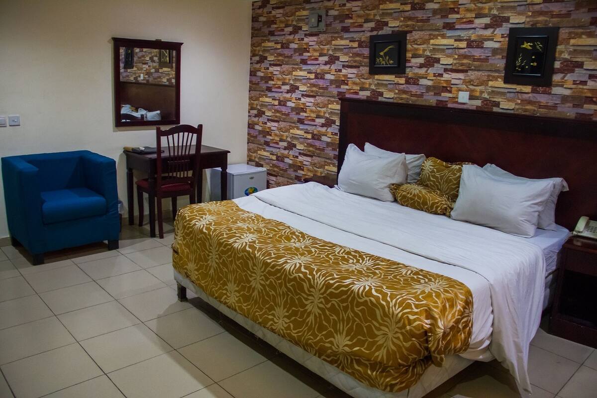 A hotel room with a bed, a blue armchair, a wooden chair, a small table, and a wall with a brick pattern.