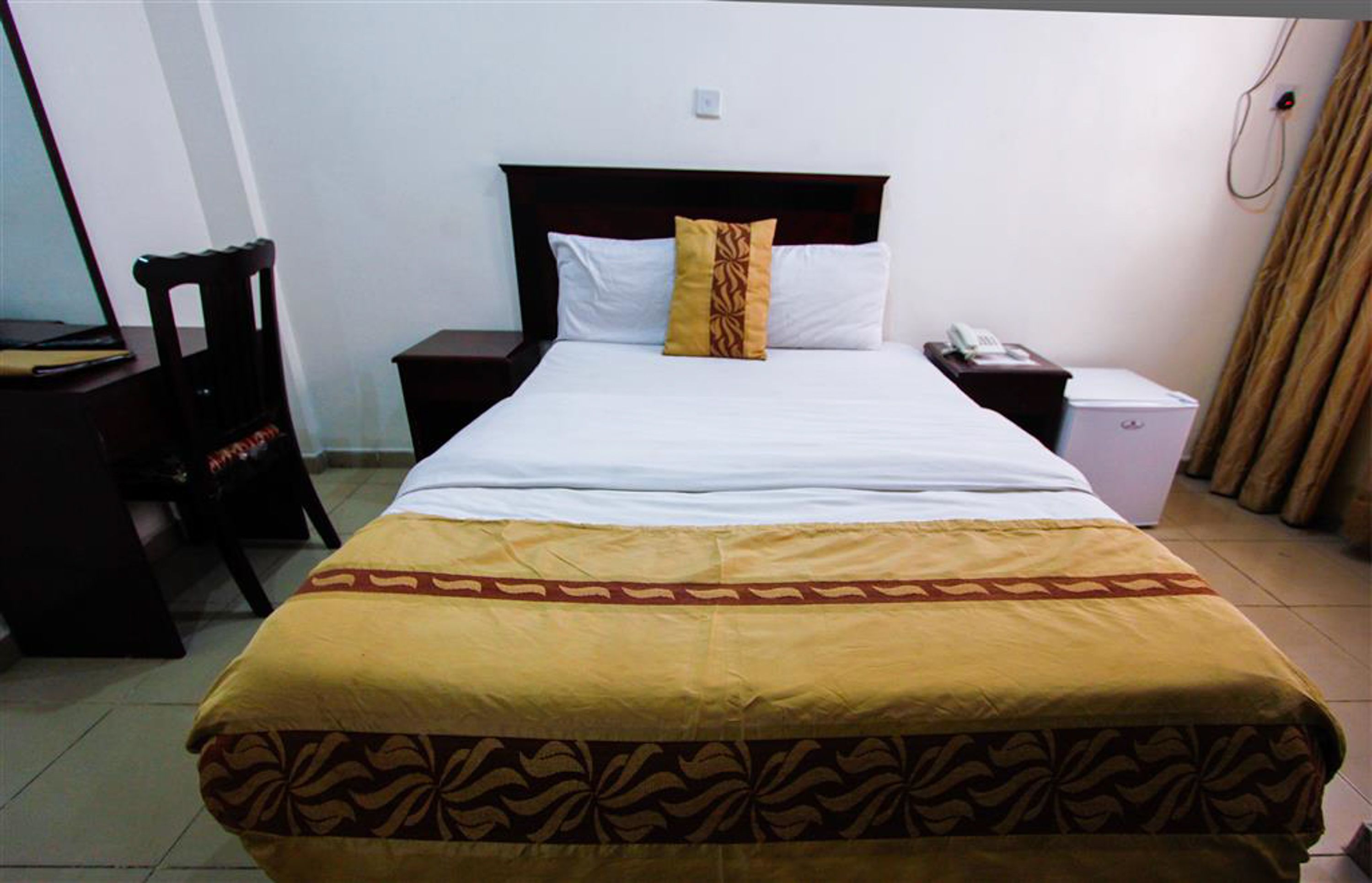 Uma cama bem arrumada com colcha estampada, uma mesinha de cabeceira com abajur e telefone.