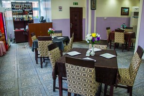 Un restaurant avec des tables dressées, des chaises recouvertes de tissus à motifs, un comptoir où sont exposées des bouteilles, et un panneau indiquant la sortie de secours sur le mur.