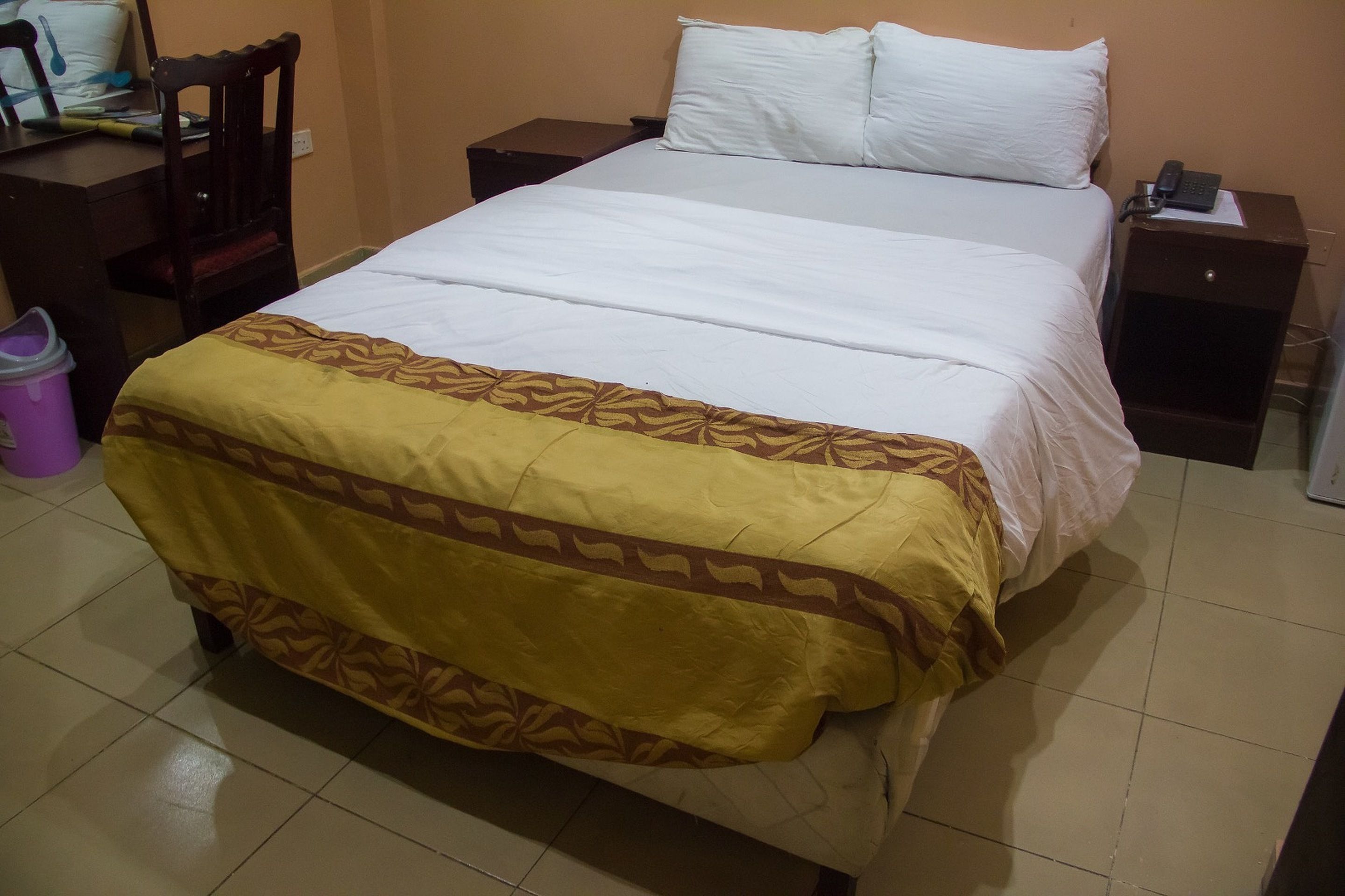 A neatly made bed with a patterned bedspread, a bedside table with a phone, and a chair with a cushion.