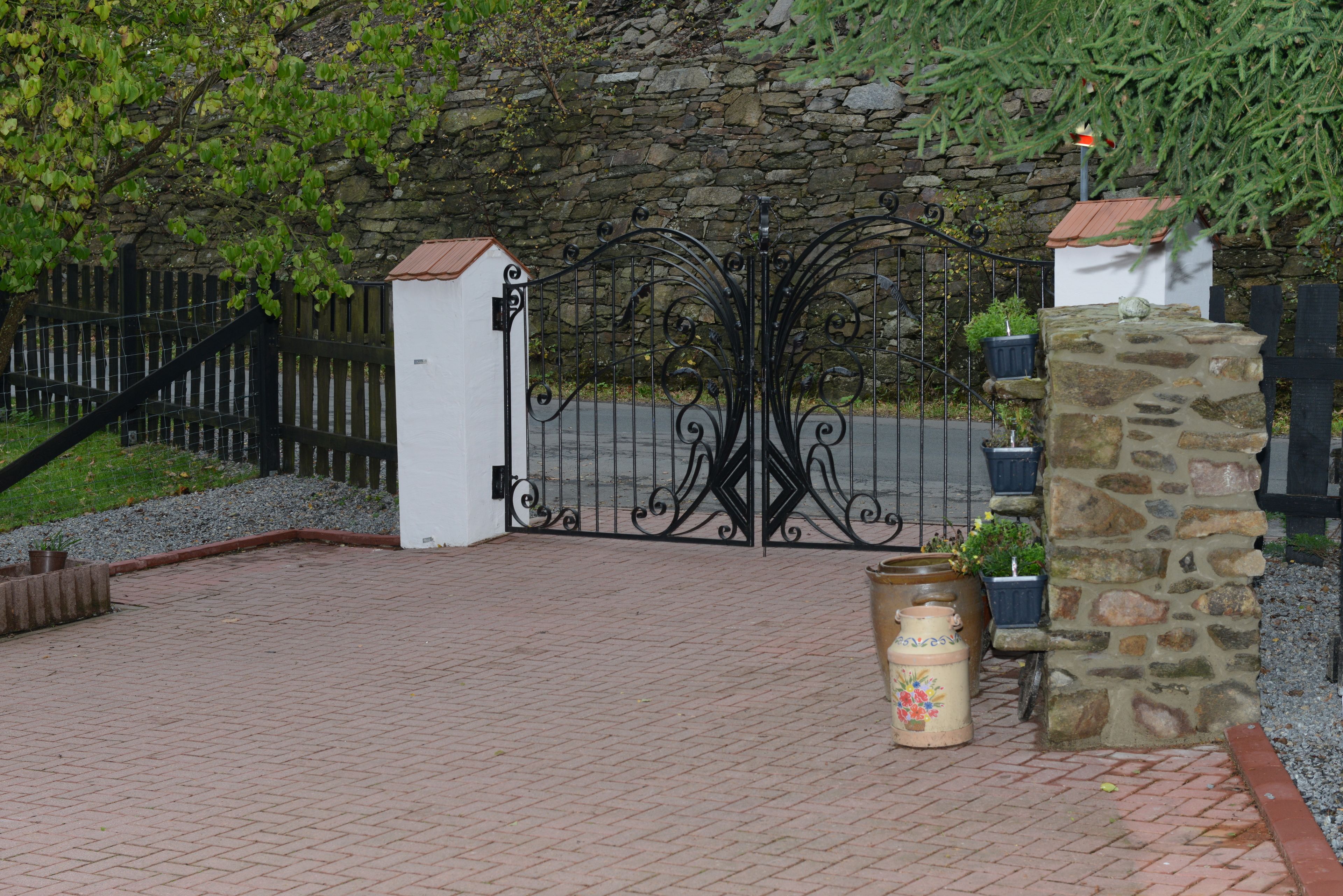 A paved entrance with a stone wall, a metal gate, and potted plants.