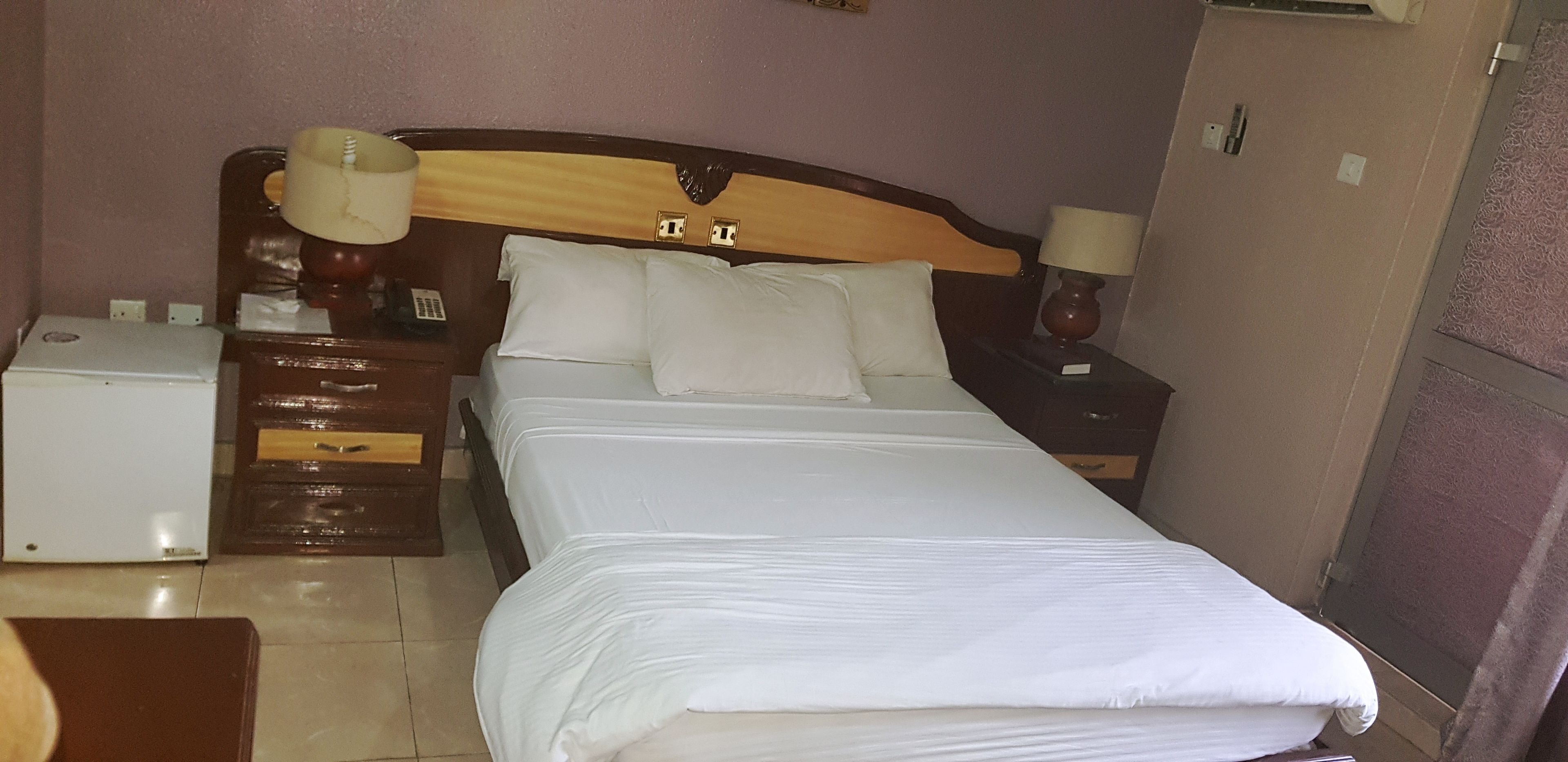A hotel room with a bed, nightstands, a lamp, a small table, and a refrigerator.