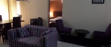 Una sala de estar con un sofá a rayas, una mesa de comedor con sillas, una mesita redonda pequeña y una cama al fondo.