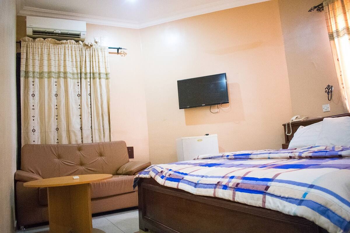 A hotel room with a bed, a sofa, a small table, a TV, and an air conditioner.