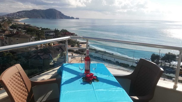 Een balkon tafeltje met een blauwe tafelkleed, een fles en een glas, met uitzicht op de kustlijn met gebouwen en een berg in de verte.