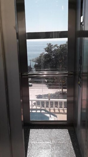 An elevator interior with a metal door, a textured floor, and a view of a street with trees and a building. - Royal Hotel Alanya (Alanya)
