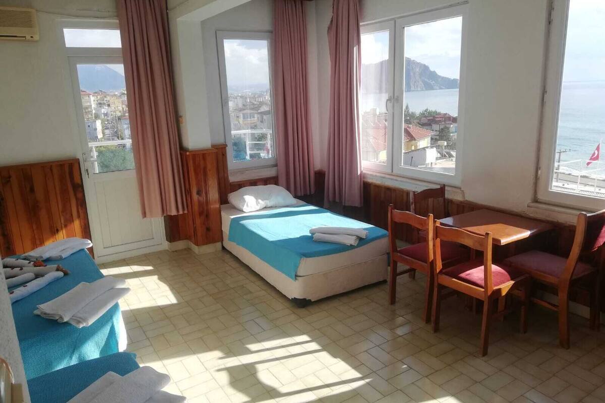 Ein Zimmer mit Bett, Tisch, Stühlen, einem Fenster mit Blick auf das Meer und Gebäude sowie einer Tür.