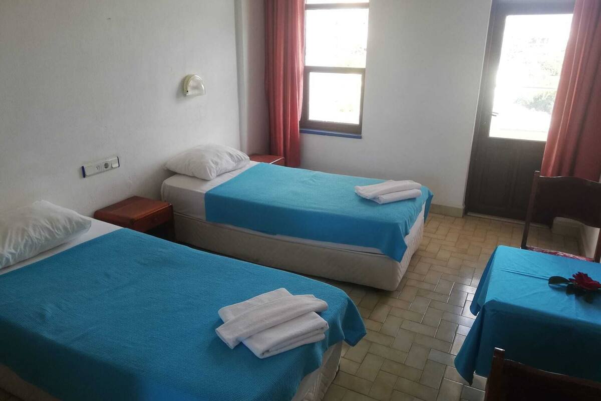 Ein Zimmer mit zwei Betten, jeweils mit blauer Bettdecke und weißen Kissen, einem kleinen Tisch mit roter Tischdecke und einem Stuhl.