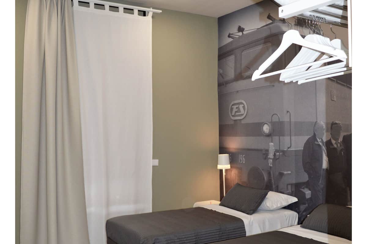 Ein Hotelzimmer mit zwei Betten, einem großen Fenster mit Vorhängen, einer Wandleuchte und einem Kleiderschrank mit Kleidung.