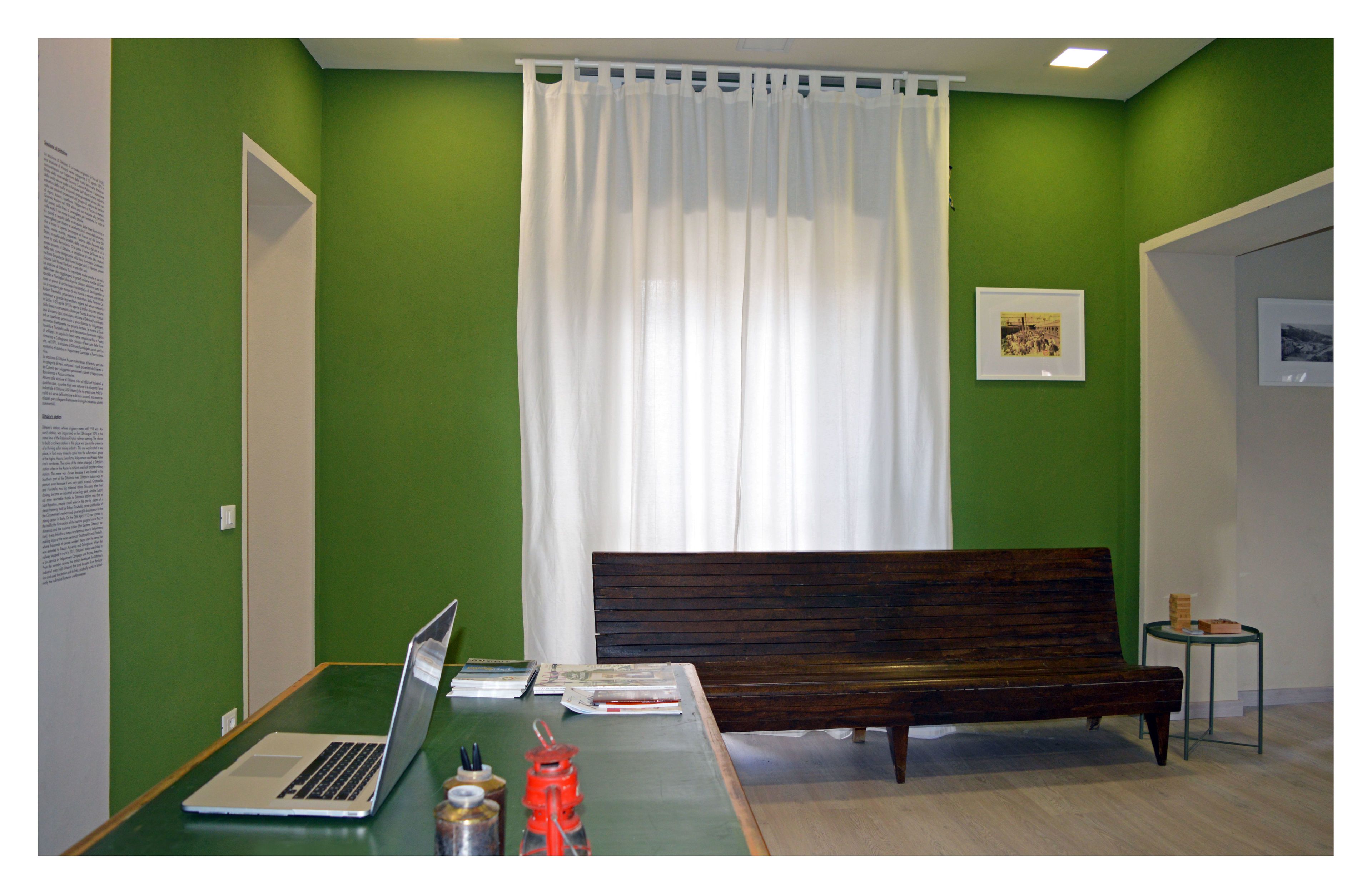 Una habitación con paredes verdes, una cortina blanca, un banco de madera, una mesa pequeña con un ordenador portátil y un cuadro en la pared.