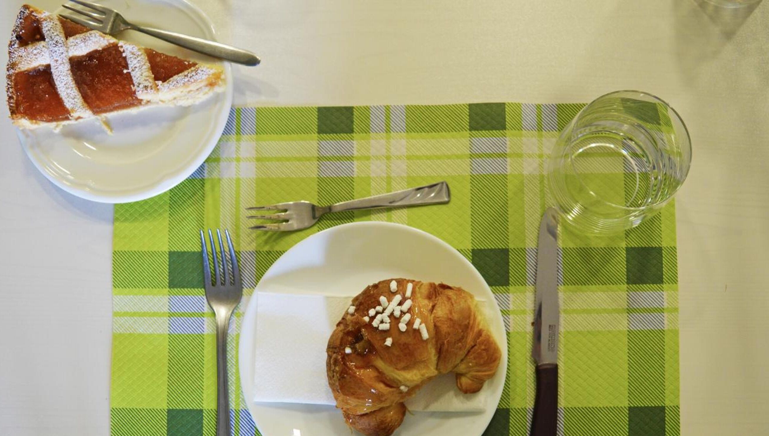 Une table avec une nappe à carreaux verts et blancs, une assiette contenant un croissant, une fourchette, un couteau et un verre d’eau.