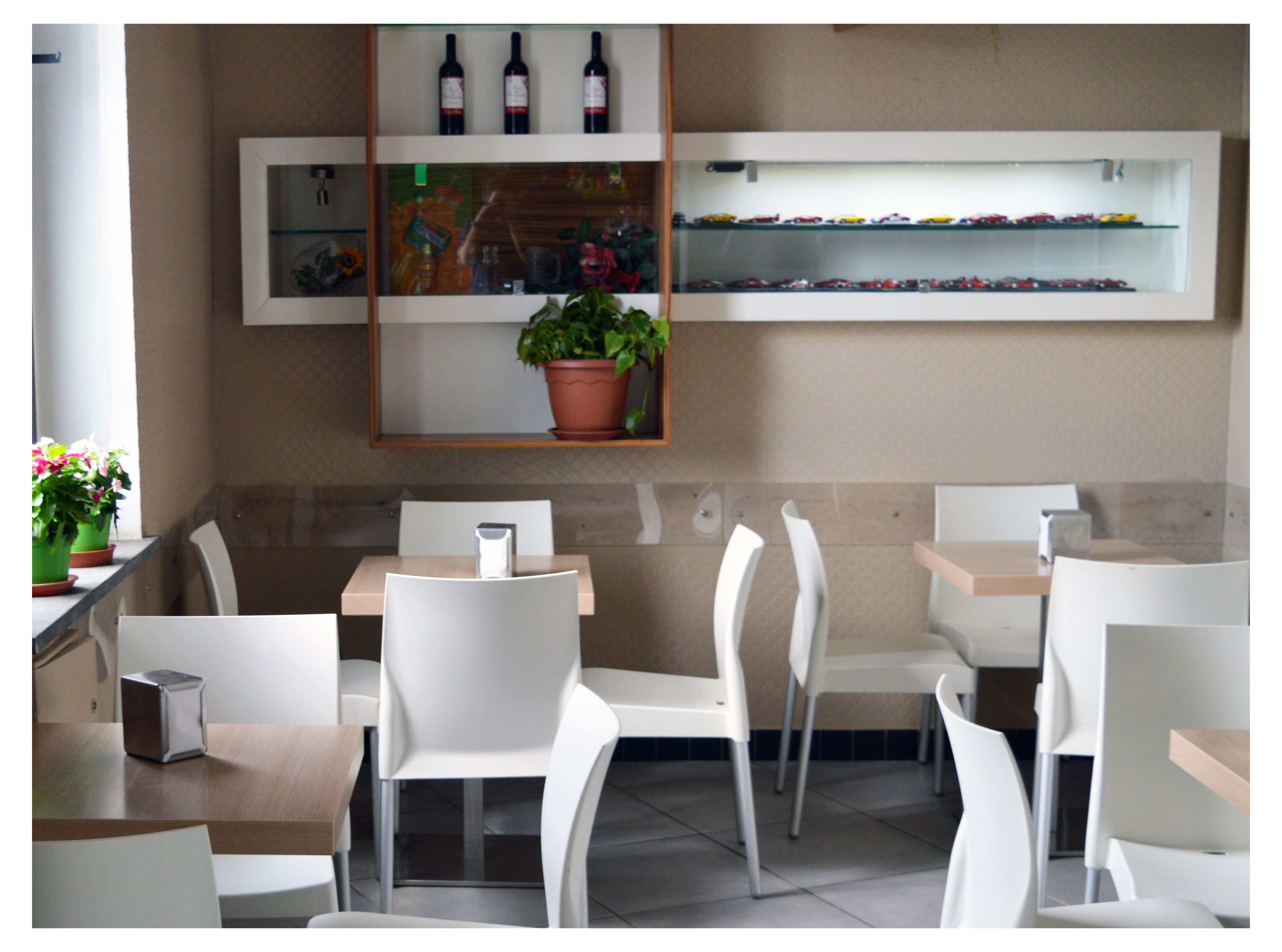 Et moderne restaurantmiljø med hvide stole og borde, en potteplante samt reoler med flasker og snacks.