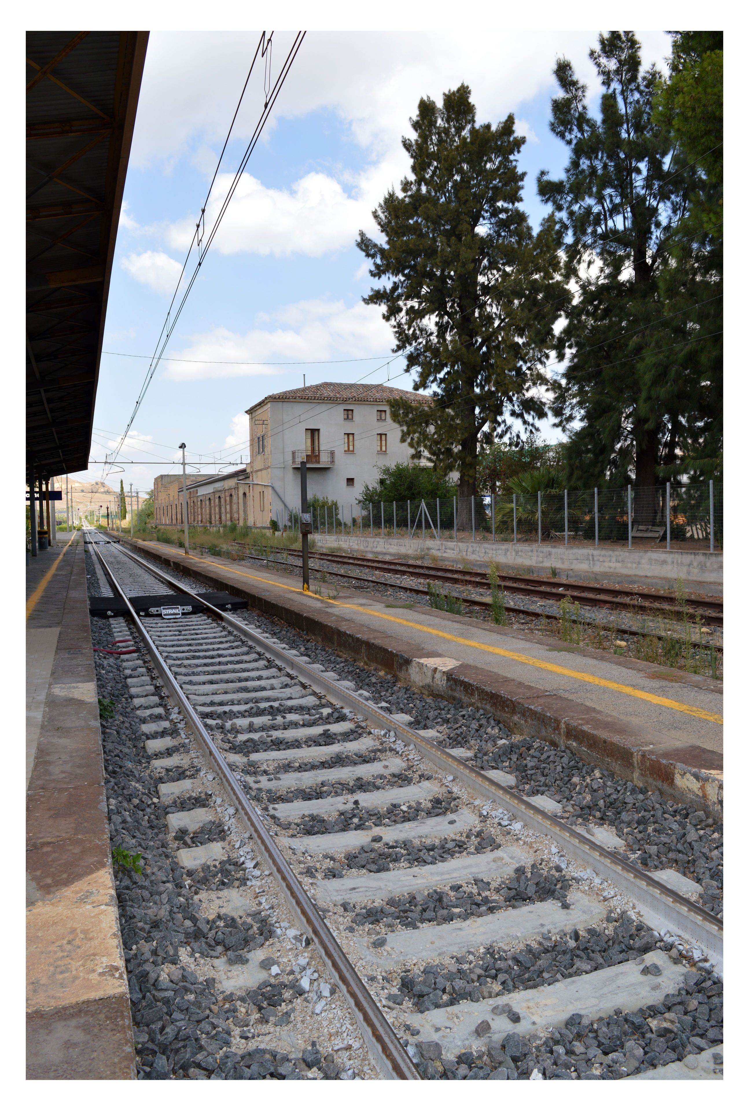 A train station platform with multiple tracks, a building with a tiled roof, and trees in the background.