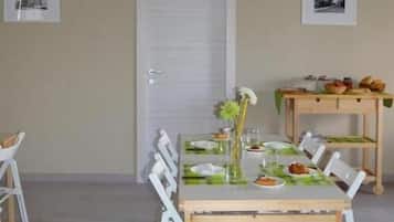 Een eetruimte met een tafel voor vier personen, stoelen, een houten bijzettafeltje met eten, en een deur met een raam.