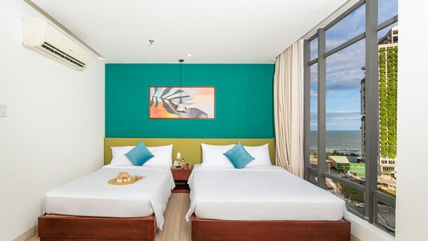 A hotel room with two beds, a teal accent wall, a large window with a view of the sea, and an air conditioning unit. - May Beach Hotel (Da Nang)
