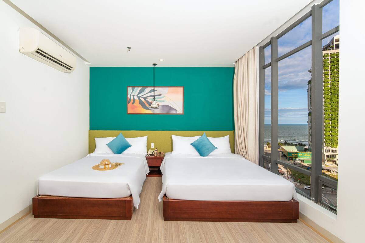 Habitación de hotel con dos camas, una pared de acento color aguamarina, un ventanal con vistas al mar y un equipo de aire acondicionado.