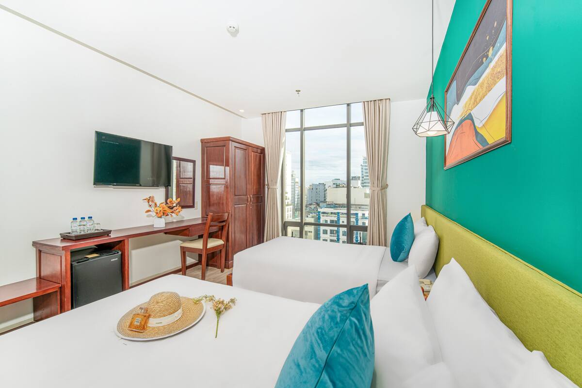 Habitación de hotel con dos camas, un escritorio con silla, televisión y vistas a la ciudad a través de amplias ventanas.