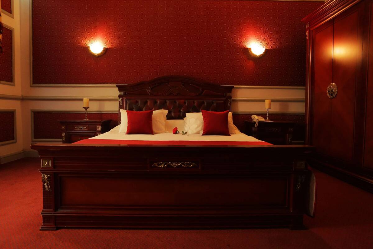 Ein großes, verziertes Bett mit Holzrahmen, ein rotes, mit Stoff bezogenes Kopfteil, zwei Nachttische mit Lampen und ein roter Teppichboden.