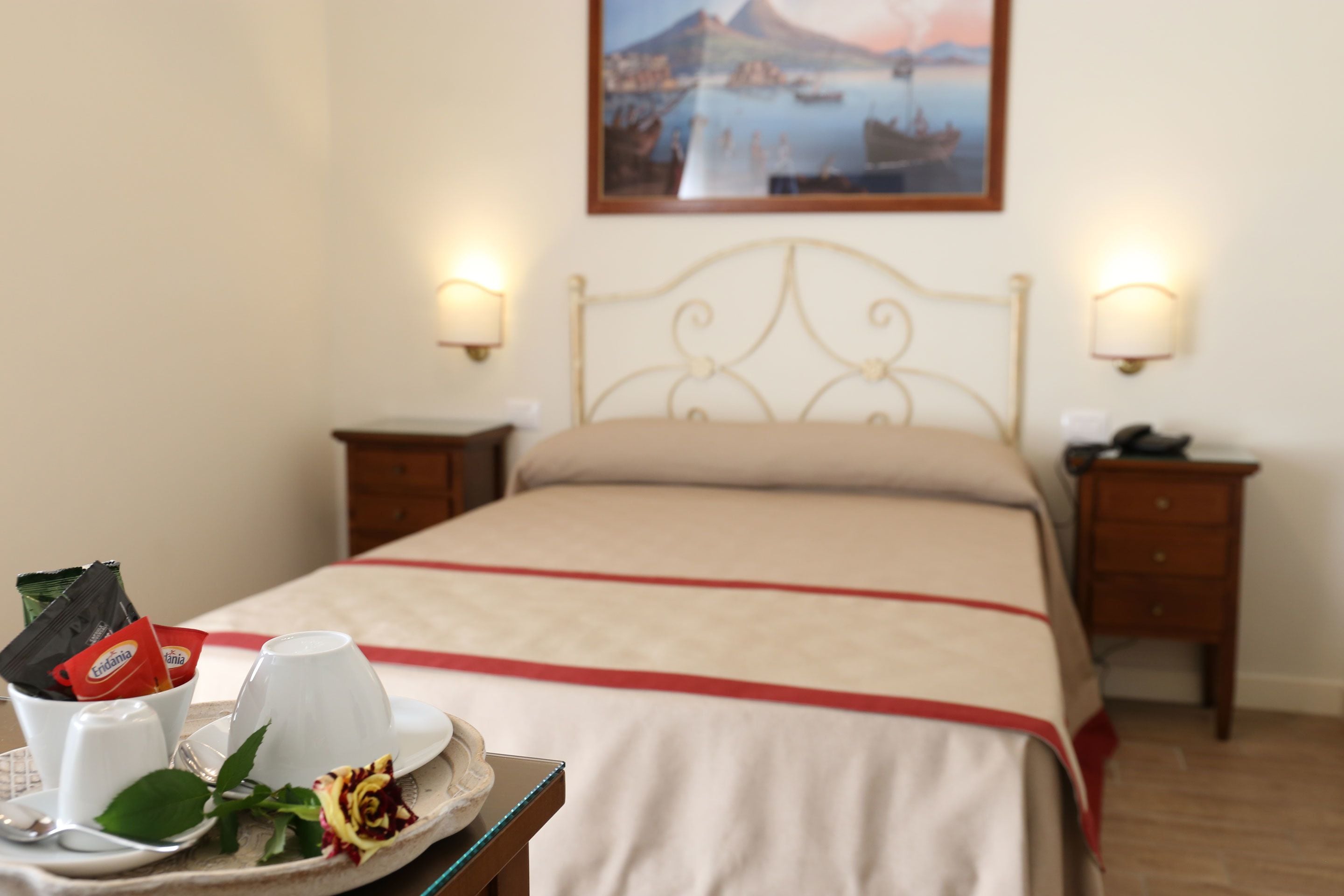 A neatly made bed with a red and white bedspread, two bedside tables with lamps, a tray with a cup, a rose, and a packet of coffee.