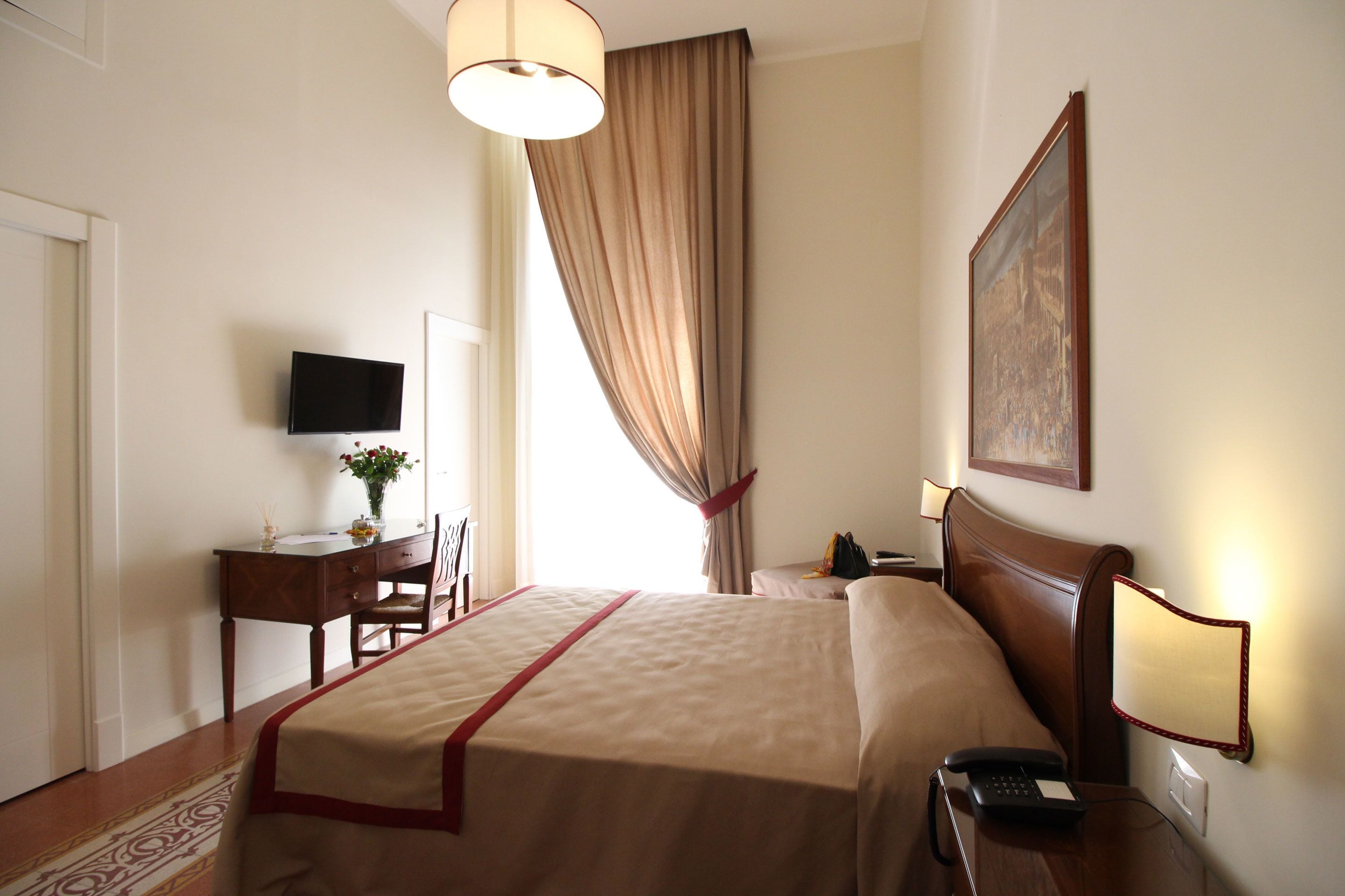 A hotel room with a bed, desk, chair, TV, and a painting on the wall.