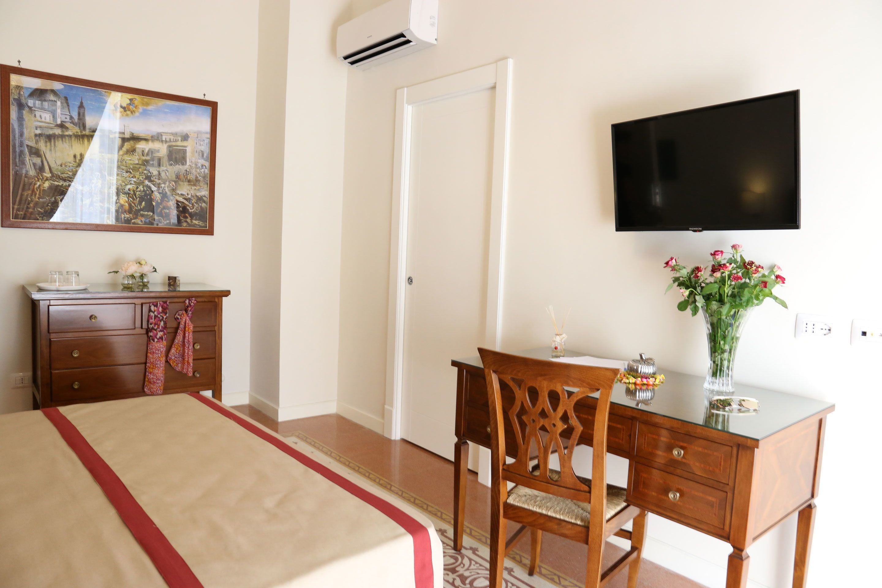 Una camera d'albergo con un letto, una scrivania in legno, una sedia, una televisione, un quadro e un vaso con fiori.
