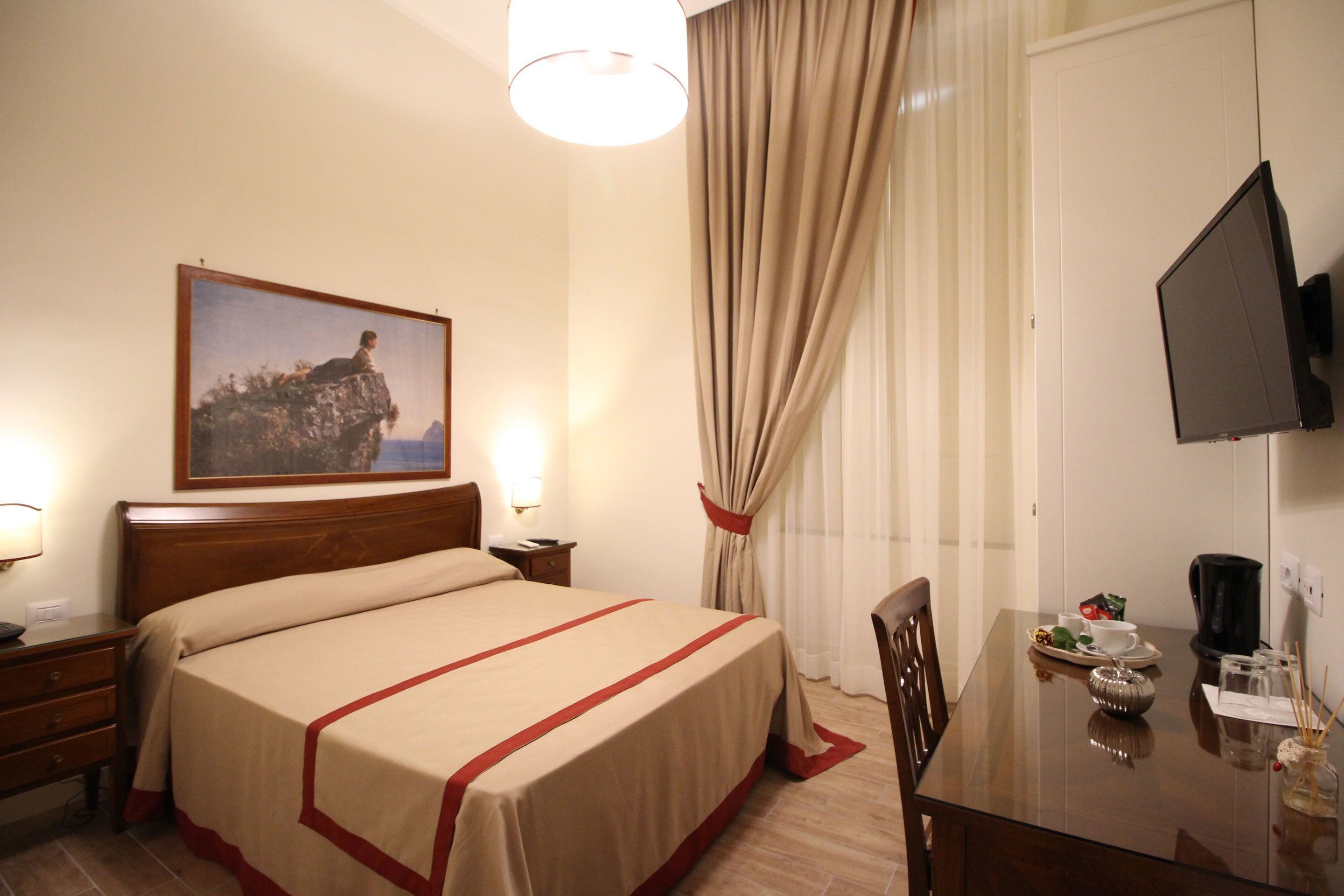 A hotel room with a bed, a desk, a chair, a television, and a painting on the wall.