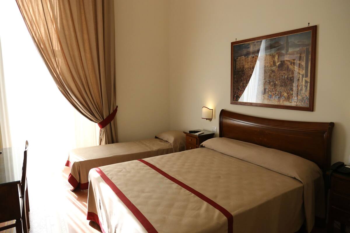 Habitación de hotel con dos camas, una mesita de noche, una lámpara, un cuadro en la pared y una ventana con cortinas.