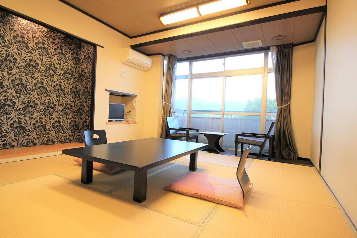 A room with tatami flooring, a low table, chairs, a TV, and a large window with curtains.