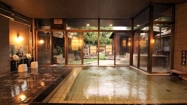 A traditional Japanese onsen with a wooden interior, a stone hot spring, and a view of the surrounding garden. - Hotel Hatta (Fuefuki)