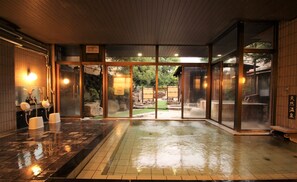 Un onsen japonais traditionnel, avec un intérieur en bois, une source d’eau chaude entourée de pierres et une vue sur le jardin environnant.