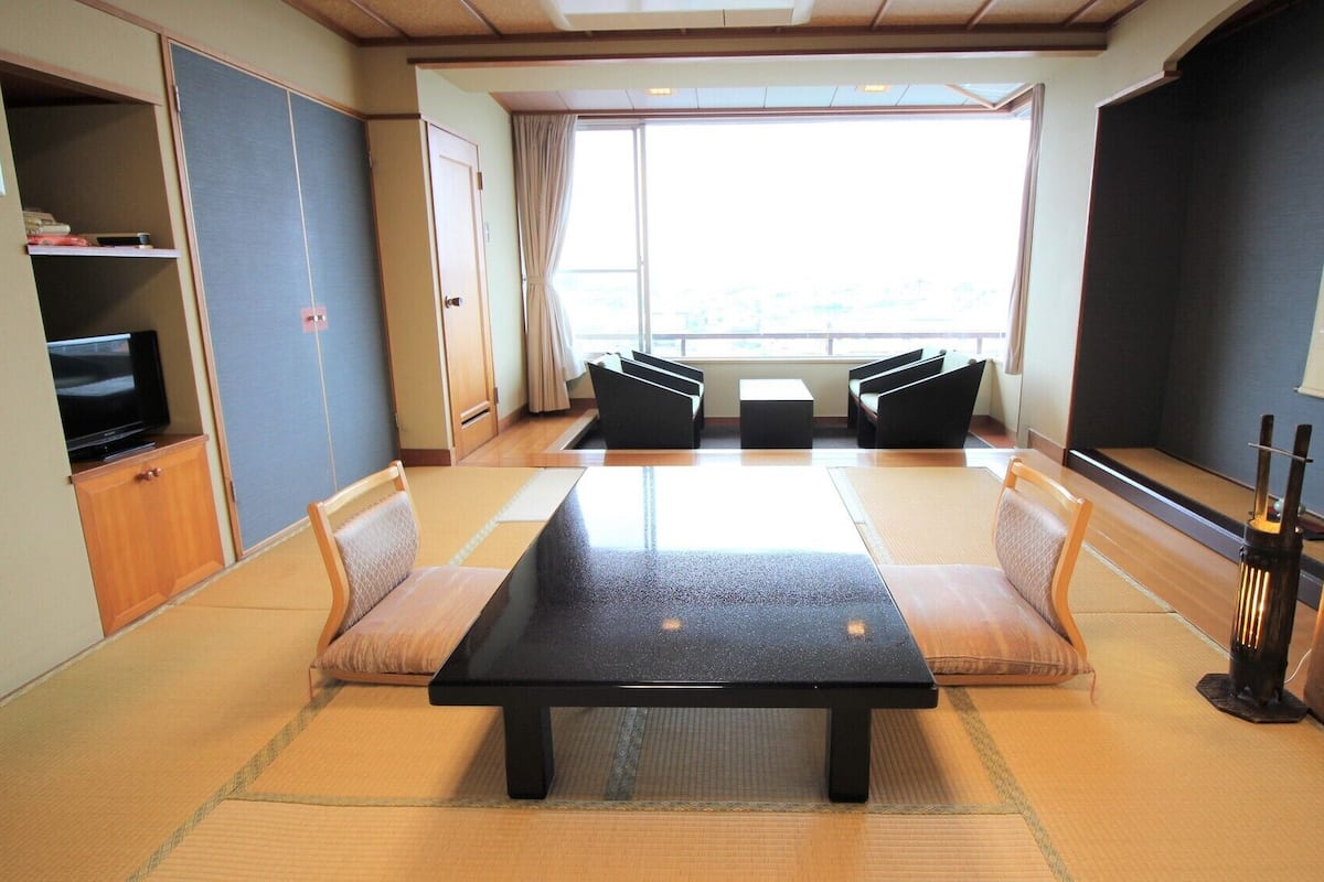 A room with tatami mats, a low table, cushioned seating, a television, and a large window.