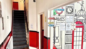 A hallway with a staircase, a chandelier, and a wall with various signs and illustrations. - Saint James Backpackers (London)