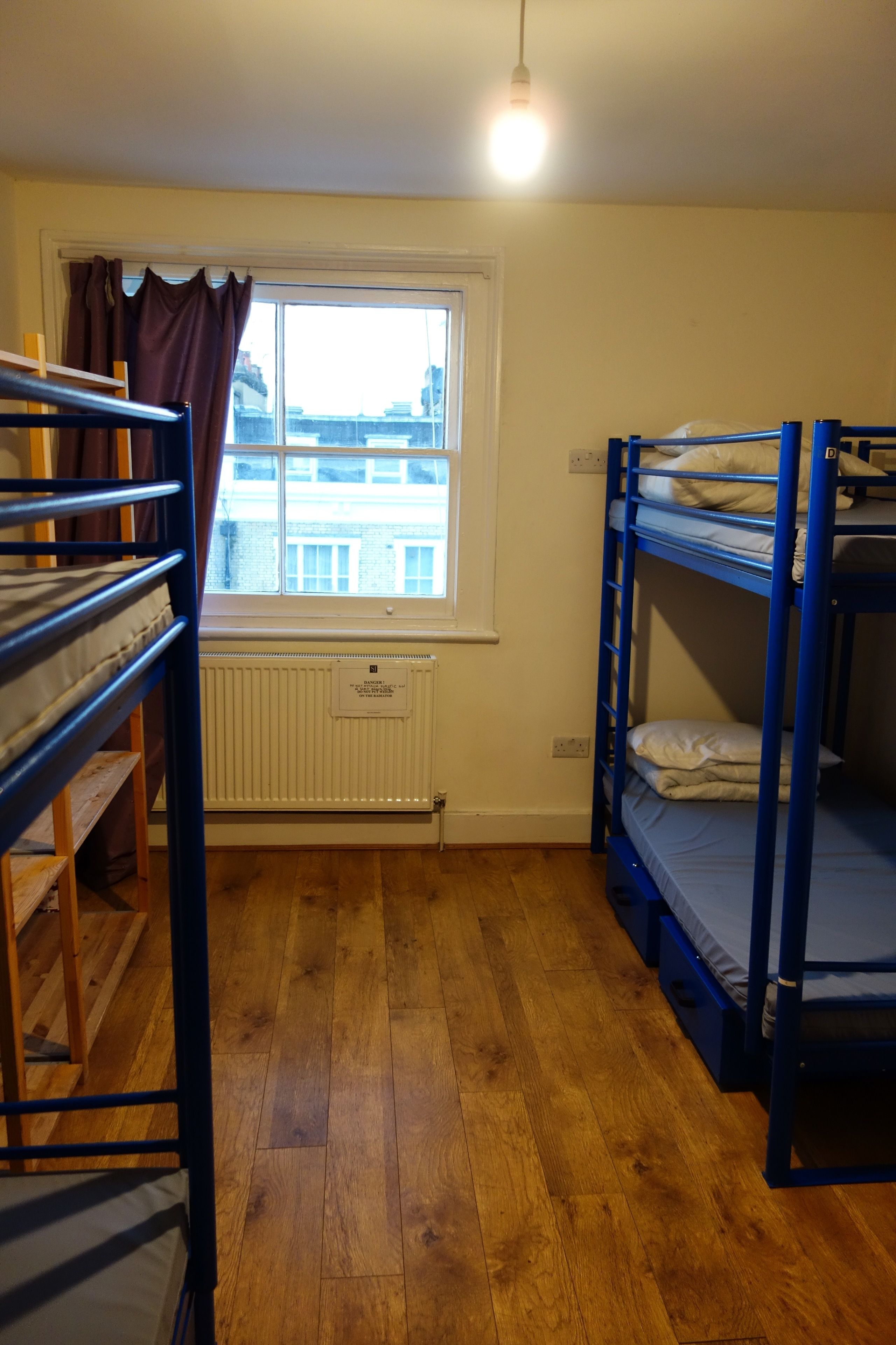 A dormitory room with bunk beds, a window with curtains, a radiator, and a wooden floor.