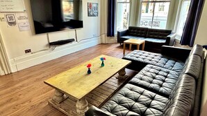 A living room with a large flat-screen TV, a wooden coffee table with vases, a leather sofa, and a small wooden table. - Saint James Backpackers (London)