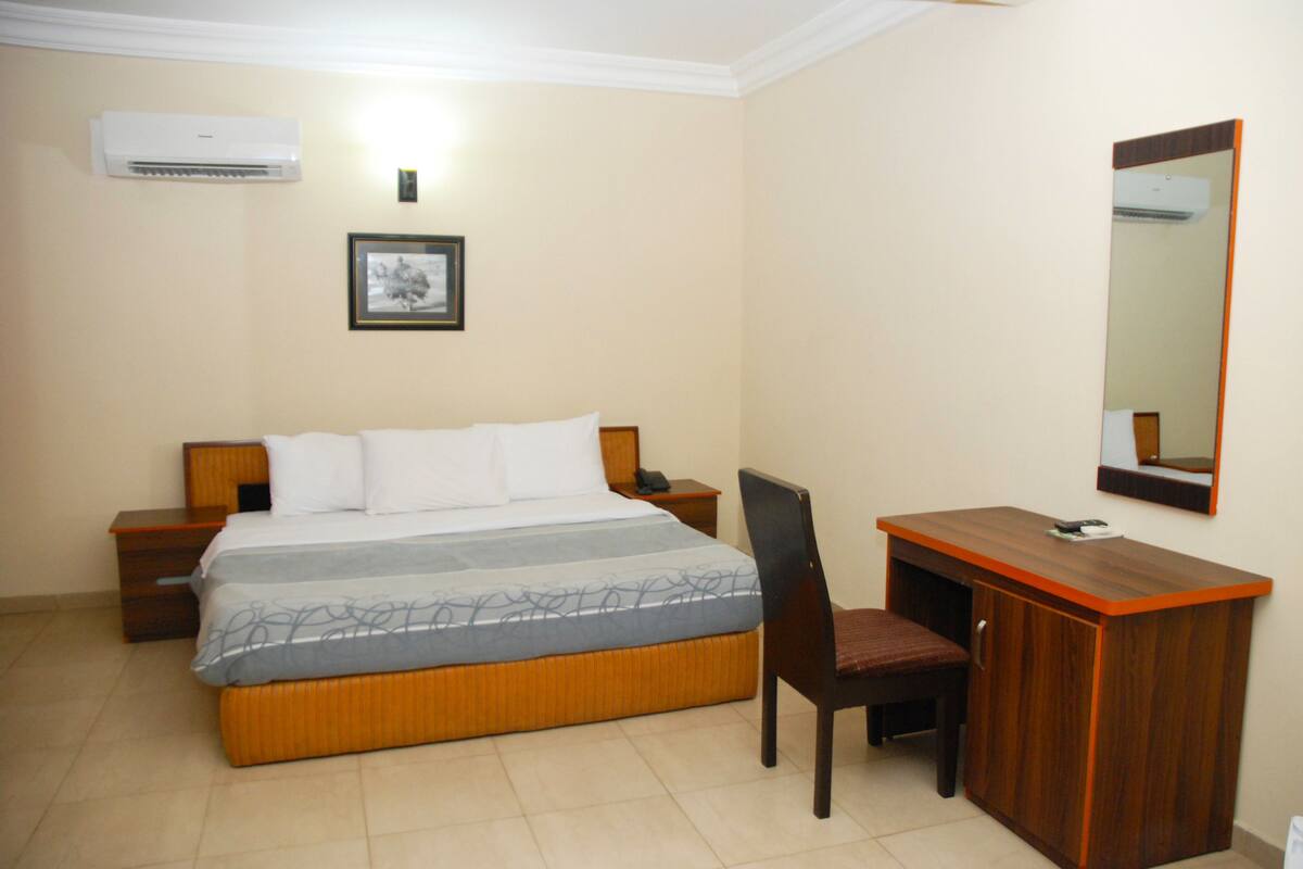 A hotel room with a bed, a desk, a chair, a nightstand, a bedside table, an air conditioner, and a framed picture on the wall.