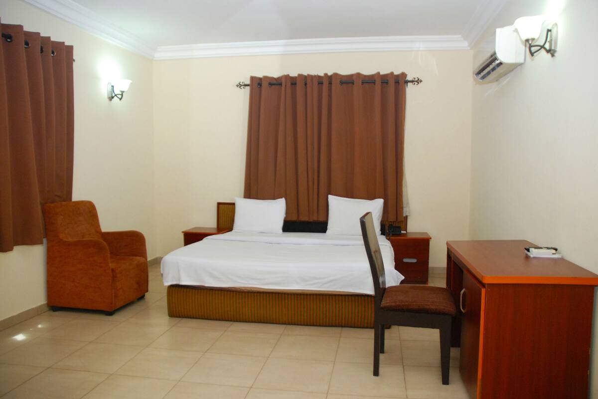 A hotel room with a bed, a chair, a desk, and an air conditioning unit.